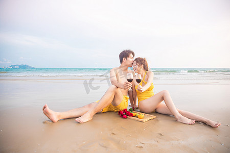 海边沙滩度蜜月摄影照片_爱情蜜月白天喝红酒的情侣沙滩捧杯亲吻摄影图配图