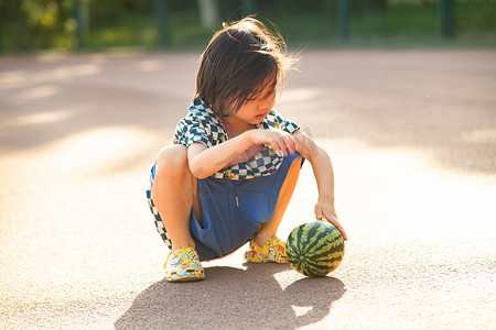 夏日儿童人像白天长发小男孩操场蹲着看西瓜摄影图配图