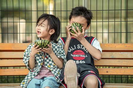 西瓜摄影照片_夏日两人人像白天兄弟二人操场长椅上开心吃西瓜摄影图配图