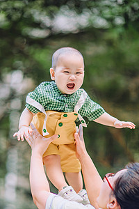 婴儿兜兜摄影照片_婴儿白天被举高高的小男孩室外举高高摄影图配图