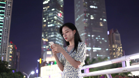 都市夜晚在高楼大厦下玩手机的年轻美女