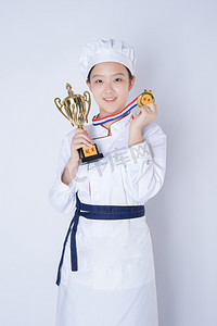 年轻女厨师白天一个女厨师白背景一手拿奖杯一手拿奖牌摄影图配图