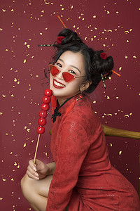国潮红金摄影照片_国潮白天穿着旗袍的美女室内吃糖葫芦摄影图配图