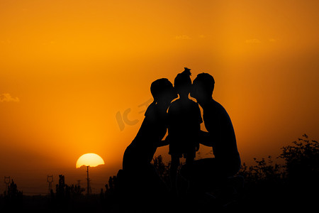 光圈摄影照片_人像剪影夕阳一家三口在夕阳下亲吻孩子摄影图配图
