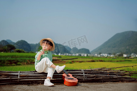 人在油菜花摄影照片_文艺清新的小男孩手拿吉他坐在油菜花田旁边摄影图配图