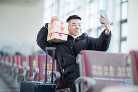 春运回家白天拿着礼物自拍的男青年候车室视频通话摄影图配图