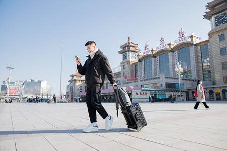 春运白天边走路边看手机的帅哥火车站拉着行李箱摄影图配图