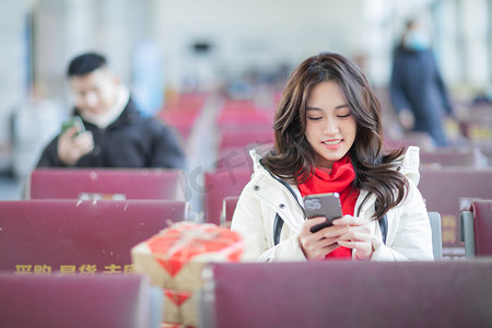 火车站人摄影照片_春运白天带红色围巾的长发美女候车室玩手机摄影图配图