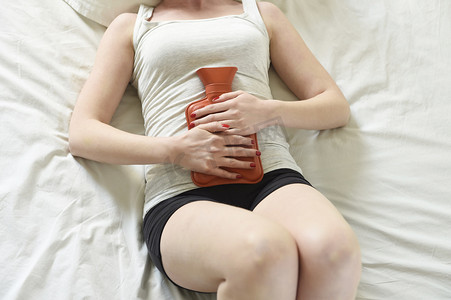 肚子疼的人摄影照片_躺在床上用热水袋捂肚子的女人