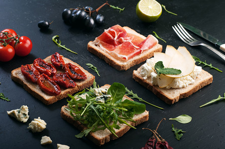 Sandwiches with a variety of toppings on a dark surface. Sandwiches with arugula, leaf mash, blue cheese, sun-dried tomatoes, slices of pear and ham. The concept of useful home cooking