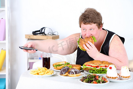 可乐炸鸡大汉堡摄影照片_拿着遥控器正在吃汉堡的胖男人