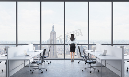 Rear view of a brunette who is looking out the window in the modern panoramic office with New York view. White tables equipped with modern laptops and black chairs.