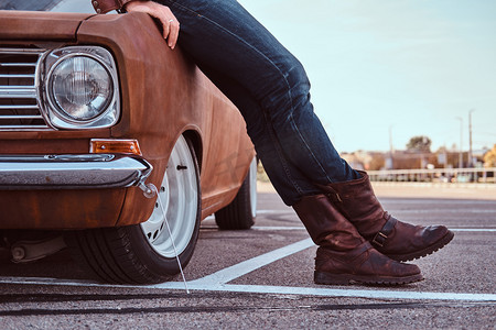 复古靴子摄影照片_男性在牛仔裤和靴子的短照片倾斜在城市停车场的复古汽车.