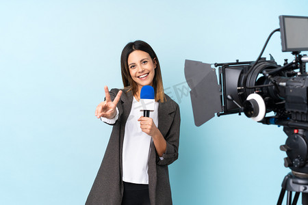 面谈摄影照片_女记者手持话筒，在孤立的蓝色背景上微笑着报道新闻，并展示胜利的标志