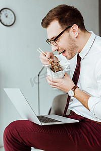 美食摄影照片_时尚的商人吃面条, 而使用笔记本电脑在办公室