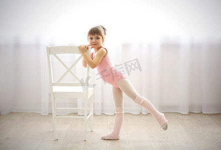 运动风格摄影照片_可爱的小女孩在舞蹈工作室制作新芭蕾运动椅边的粉红色紧身连衣裤
