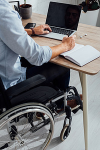 残疾人摄影照片_坐在轮椅上的男子在笔记本上写字和在办公桌旁使用笔记本电脑的剪影