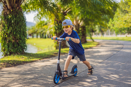 滑板男孩摄影照片_孩子们在公园里踢摩托车。孩子们学习滑板。在阳光明媚的夏日里滑冰的小男孩。儿童在安全居住街道上的户外活动。学龄前儿童的积极运动