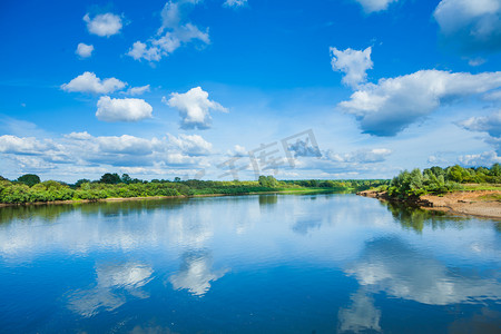 宽阔的河流与反射和绿色布什对海岸和蓝色多云的天空