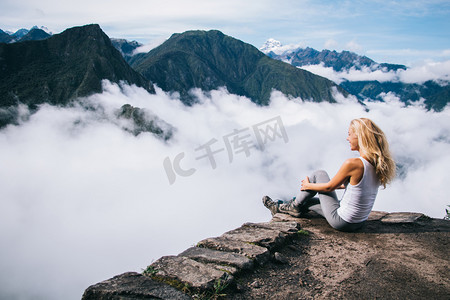 惊险摄影照片_正值年轻的金发女郎穿着积极的穿着坐在洛基山和欣赏惊险的风景高绿色的山覆盖在南美惊人的旅程中的白色雾