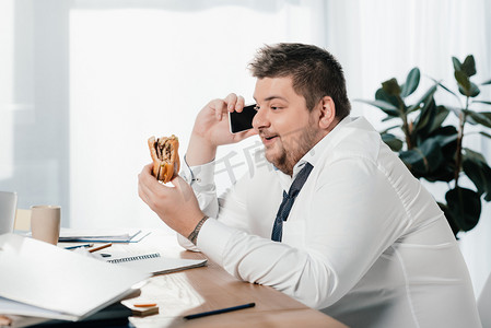 在办公室吃汉堡包的时候, 超重的生意人在谈论智能手机