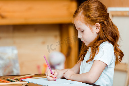 可爱的红头发小孩拿着彩笔在纸上画画 