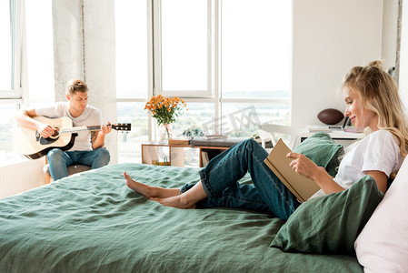 年轻的妇女在床上看书, 而男友在家里玩声学吉他