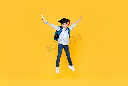 毕业跳摄影照片_英俊的男生，戴着研究生帽，笑着跳到黄色的背景上学习教育理念