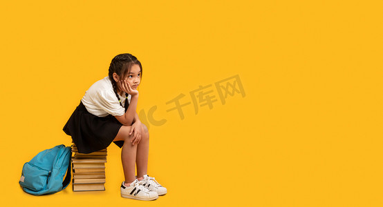 坐着拍背摄影照片_不快乐的女学生坐在书架上，黄色背景，全景