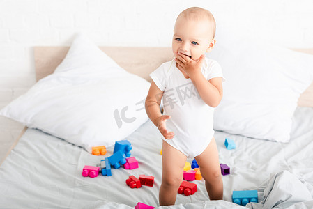 微笑赤脚婴儿在白色的衣服站在床上与玩具，并采取手指到他的嘴里