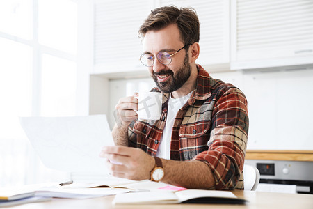学习规划表摄影照片_年轻留着胡子的男人戴着眼镜边喝茶边在家里做笔记的画像