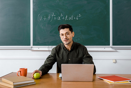坐在电脑桌前、拿着苹果、在教室里看相机的男老师