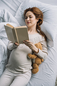 抱抱摄影照片_美丽孕妇抱着泰迪熊和卧室看书的高角度观