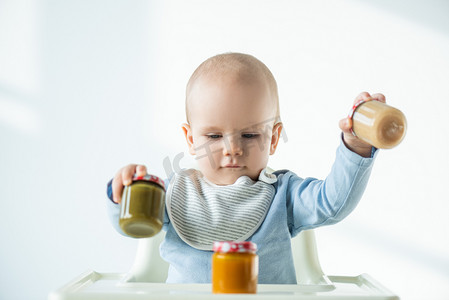 vegetable摄影照片_Selective focus of baby holding jars of vegetable baby nutrition while sitting on feeding chair on white background