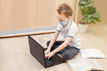 家庭远程在线学习。一个戴防护面具的孩子坐在地板上，在电脑上工作。那个学生在做作业.隔离。Coronavirus 。呆在家里。家庭教育。小心点.