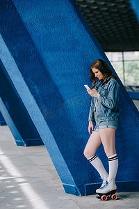 欧美场景摄影照片_时尚女性在耳机, 牛仔服装, 高袜子和复古轮滑鞋使用智能手机的侧面视图