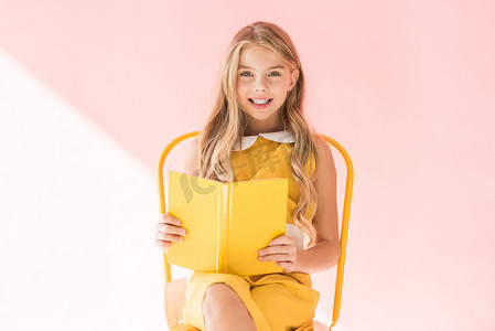 微笑时尚的孩子看书而坐在黄色的椅子上粉红色