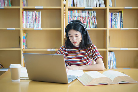 青少年人物摄影照片_一个女大学生坐在桌旁做大学图书馆的作业。 学生使用笔记本电脑，在网上学习.