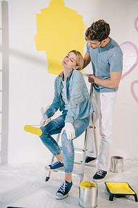 一个英俊的男人站在疲惫的女朋友旁边，坐在梯子上，手里拿着黄色的油漆滚筒