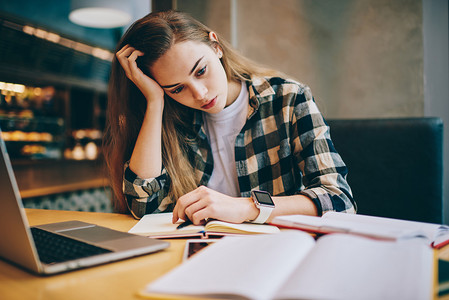 疲惫的女学生准备重要的考试, 试图在室内使用现代笔记本电脑和书籍找到问题的答案, 集中的女性从大学图书馆的课程工作中感到头疼