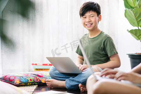 坐地板上摄影照片_聪明的亚洲少年使用电脑笔记本电脑，坐在木制地板上，笑着，与朋友讨论小组工作、家庭作业、考试准备等问题。小组工作与协作.