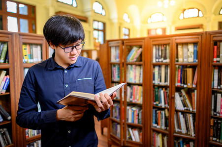 阅读看书学习摄影照片_聪明的亚洲人大学生戴着眼镜，坐在老式书架上看书。高校图书馆的教材资源用于教育科目和研究.教育机会奖学金.