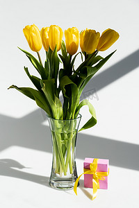 母亲节活动活动摄影照片_在白色的母亲节概念的礼品盒旁边花瓶里的黄色郁金香上的阳光