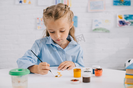 portrait of adorable focused child drawing picture with paints and brush at table