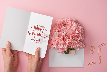 装饰文字装饰摄影照片_妇女拿着贺卡与愉快的妇女天的文字在康乃馨花附近的裁剪视图在信封在粉红色的背景