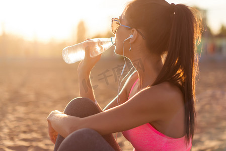 beber摄影照片_户外锻炼在傍晚夕阳夏天在海滩室外肖像上下班后美丽健身运动员女人喝水.