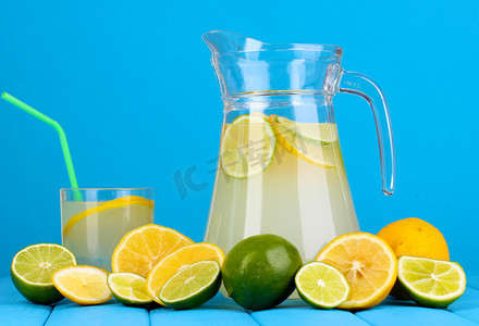 柠檬汁背景摄影照片_柑橘柠檬汁在投手和柑桔围绕在蓝色背景上的木桌上的玻璃
