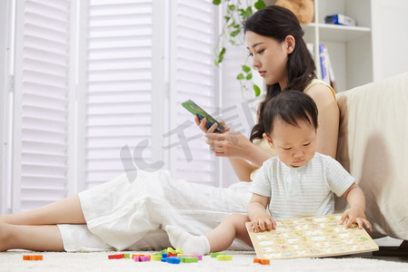 成年人玩具摄影照片_陪着宝宝玩耍的年轻妈妈使用手机