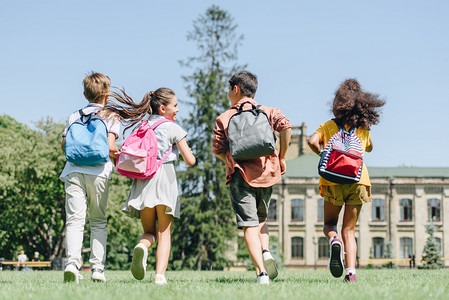 back view of four multiethnic schoolkids with backpacks running on lawn in park