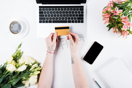 cropped view of woman holding credit card near laptop, smartphone with blank screen, cup of coffee and bouquets on white